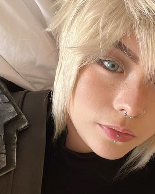 A close-up selfie of a person with platinum blonde hair, light blue-green eyes enhanced by Like Me Tide Blue lenses, and edgy piercings, set against a white pillow and dark clothing with metallic details.