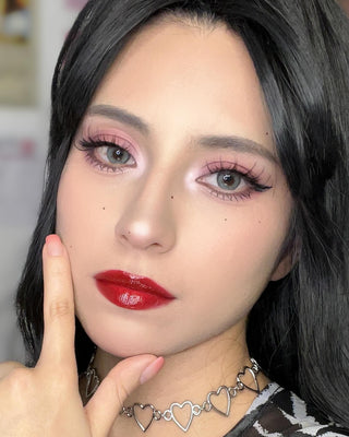 A close-up of a person with sleek black hair wearing Moonbeam Navy Gray contact lenses, bold red lipstick, soft pink eyeshadow, and fluttery lashes. They pose with one hand near their face, accessorized with a silver heart-chain necklace, exuding a glamorous and chic vibe.