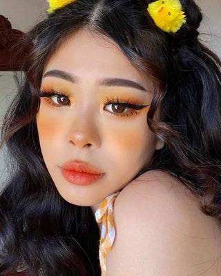 A close-up of a woman with long, wavy hair wearing Neo Dali Extra Brown contact lenses. Her makeup features bold yellow-orange eyeshadow, dramatic lashes, and soft orange lipstick. She accessorizes with playful yellow chick hair clips, complementing her cheerful, summery look.